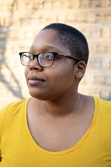 Deidra is visible from the chest up. She wears thick-rimmed dark brown glasses, small round gold earrings, and a yellow-gold short-sleeved t-shirt, her black hair cut close to her head. She looks up and to her right, standing in front of a wall of large beige bricks that has shadows cast upon it.